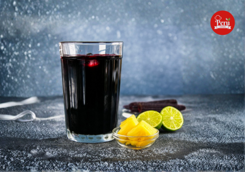 vaso de chicha morada peruana con limón verde y trozos de piña al lado sobre una mesa.